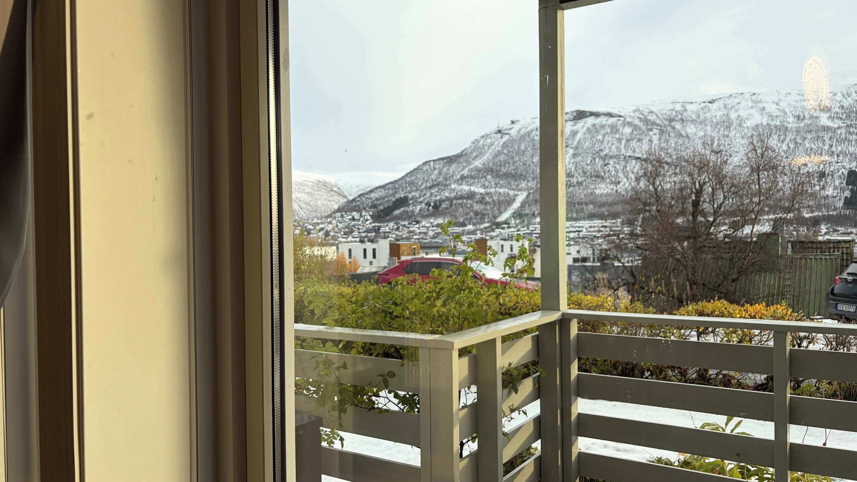 Un balcon avec vue sur une montagne enneigée, une voiture rouge et un bâtiment en arrière-plan.