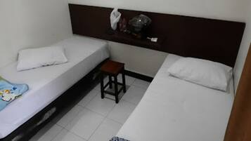 Una habitación con dos camas, un taburete pequeño, una estantería con objetos y una pared blanca.