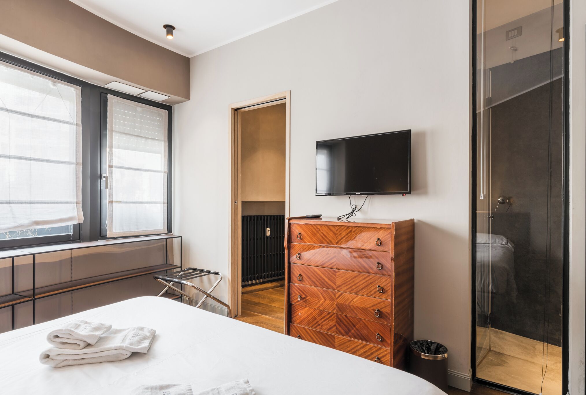 A modern hotel room with a bed, a wooden dresser, a TV, and a shower.
