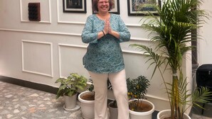 A person standing in a hallway with framed artworks on the wall, potted plants, and a tiled floor. - House Of Amer (Amer)