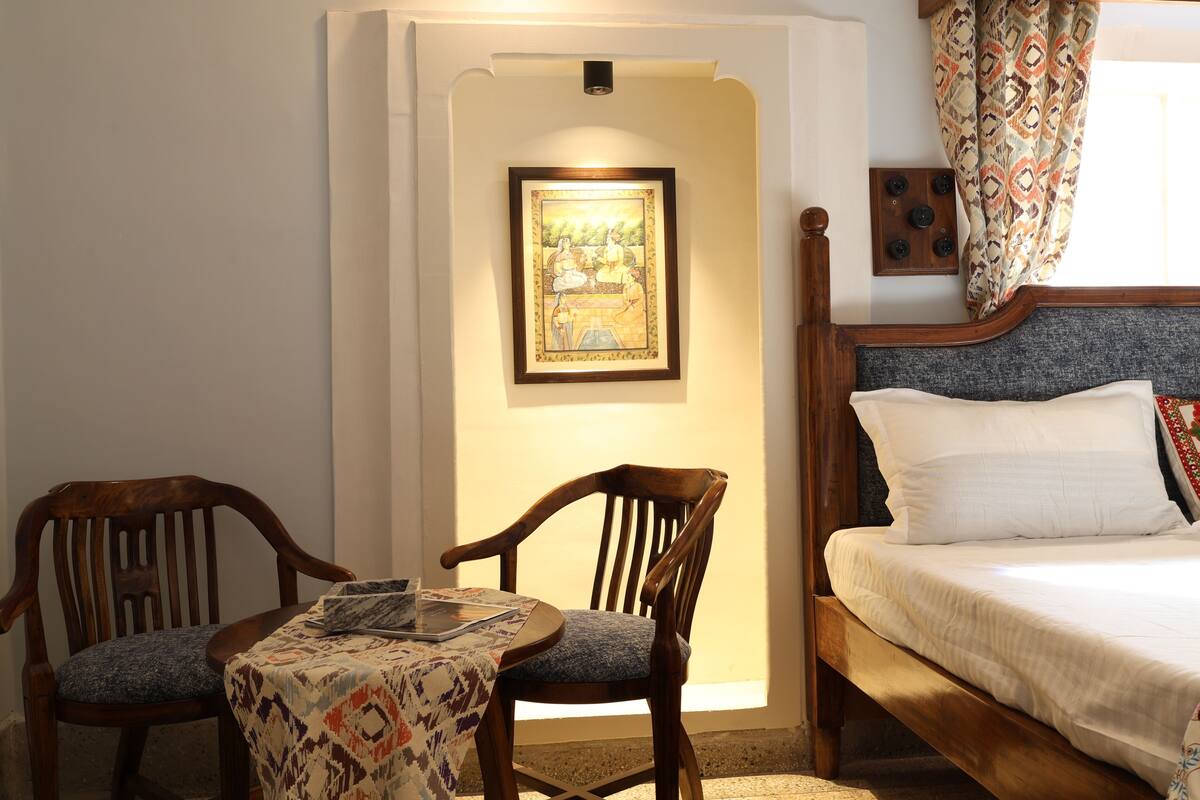 Un dormitorio con una cama de madera, dos sillas de madera, una mesita con mantel estampado y un cuadro enmarcado en la pared.