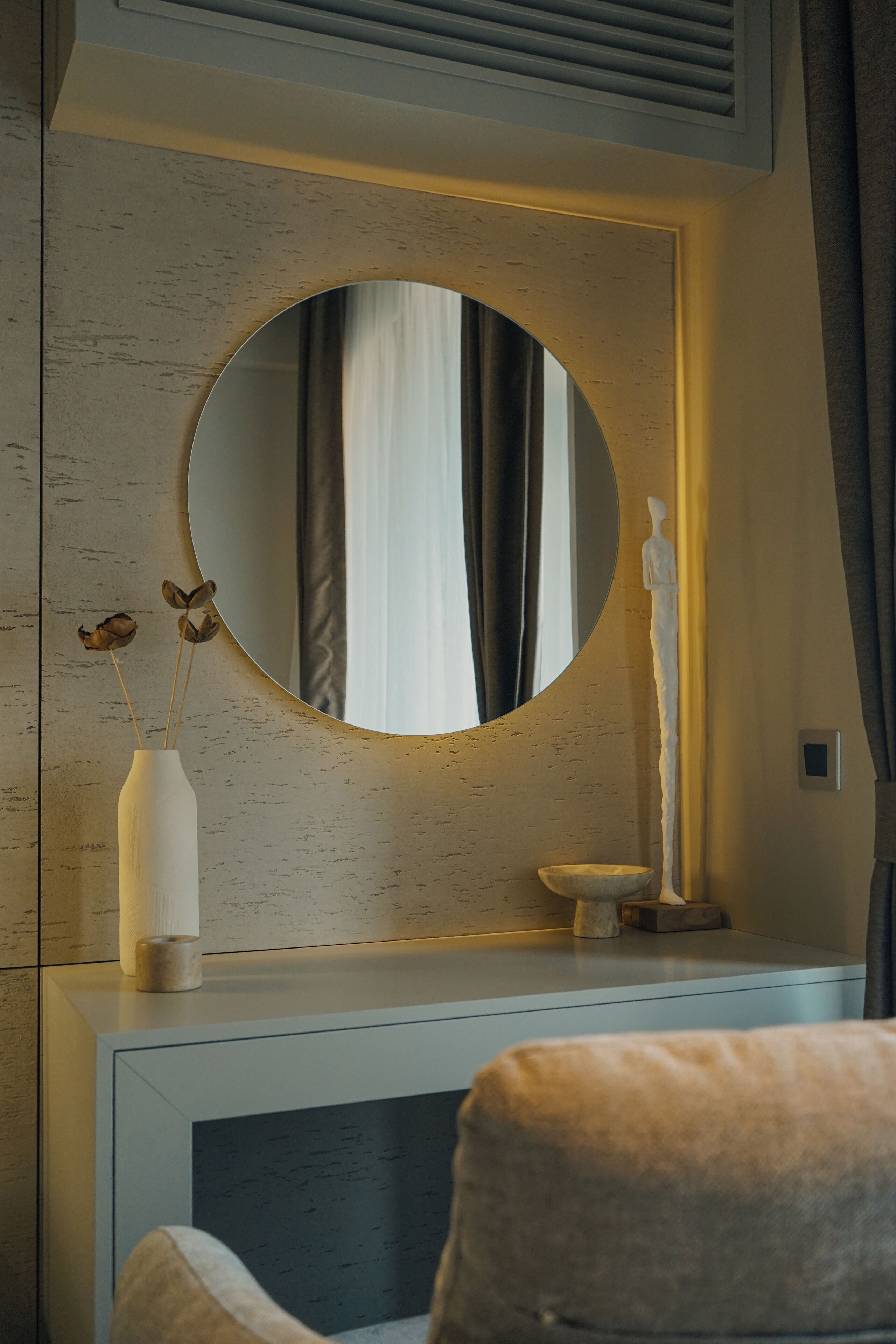 A modern bathroom with a round mirror, a vase with dried flowers, and a statue on a pedestal.