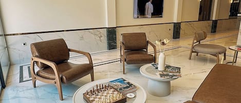 A lobby with marble flooring, leather armchairs, a chess table, and a television screen.