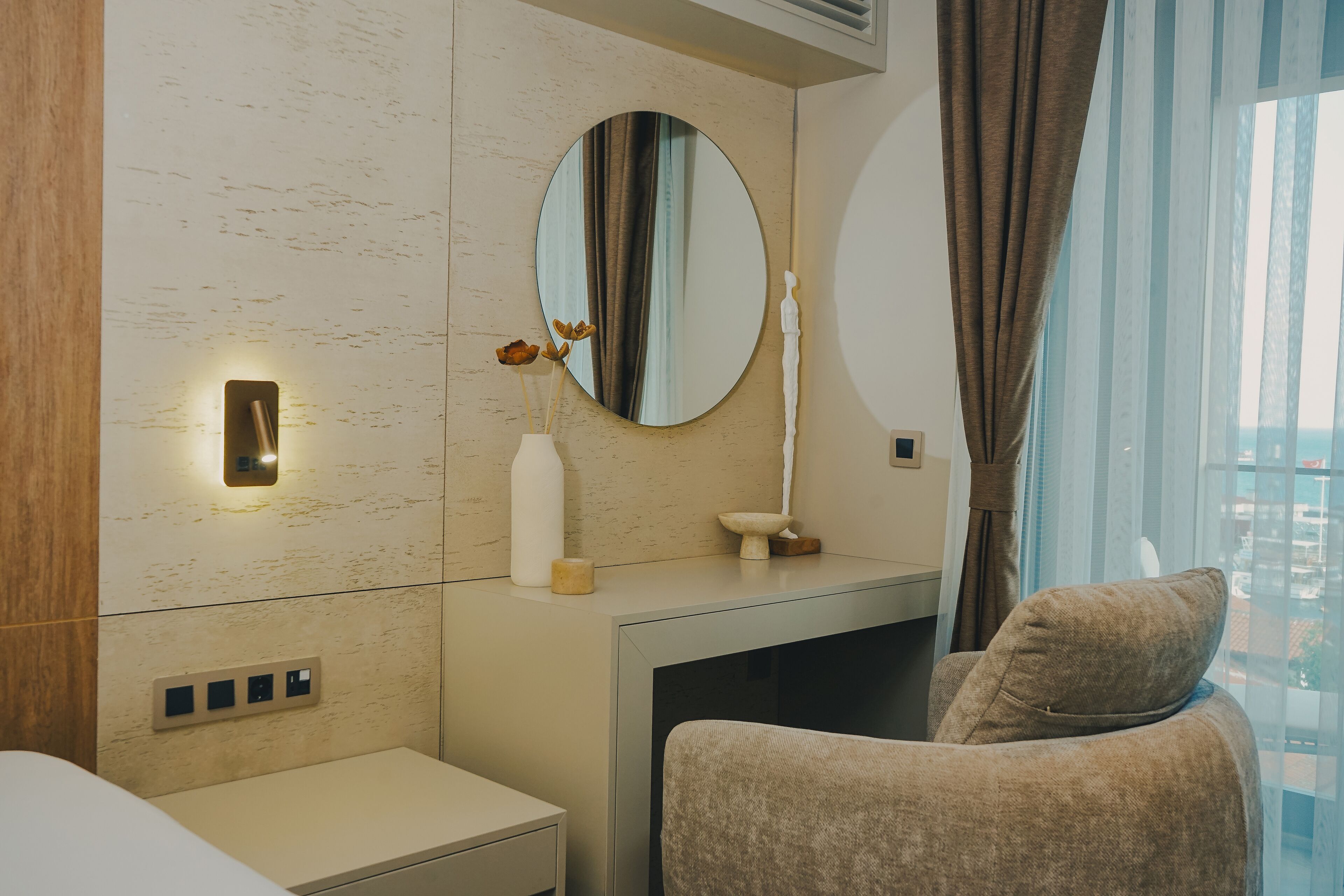 Et moderne hotelværelse med minimalistisk design, der indeholder et stort spejl, et natbord med en vase blomster og et behageligt siddeområde med pude.