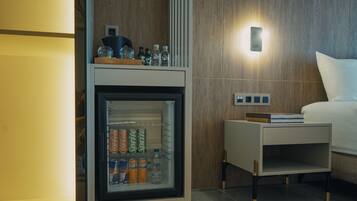 A minibar with a refrigerator containing beverages, a bedside table with a book, and a wall-mounted light.