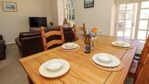 Uma sala de jantar com uma mesa de madeira posta para quatro pessoas, uma televisão ao fundo e uma janela com vista para o exterior.