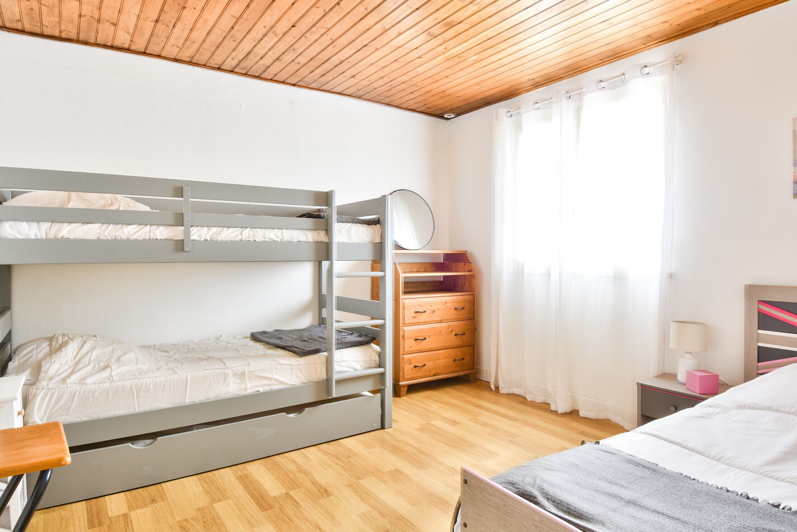 Et soveværelse med køjeseng, en trækommode, et natbord med lampe og et vindue med gardiner.