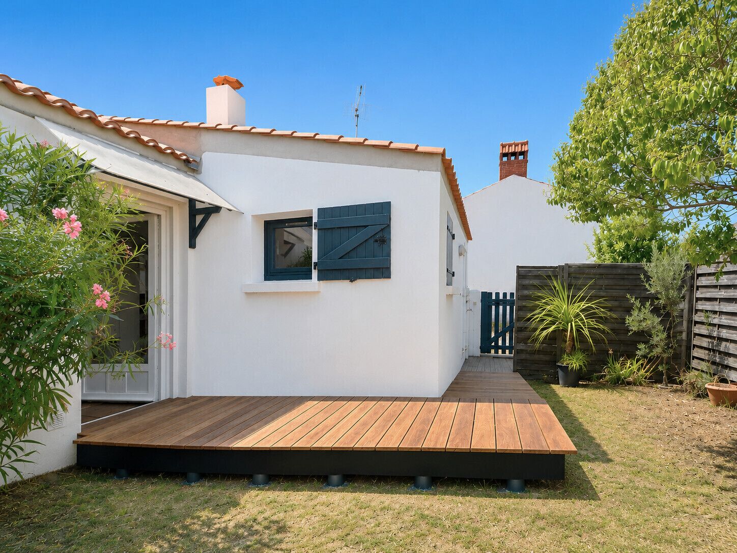 Un edificio bianco con un patio in legno, una finestra con persiane e un piccolo giardino con piante.