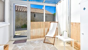 Un patio iluminado por el sol con una silla blanca, una mesita con un hervidor y vista a un edificio de piedra a través de una puerta de cristal.