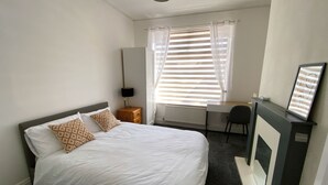 A hotel room with a bed, a desk, a chair, a radiator, and a window with curtains. - Modern room for short stay in Salford
ONLY SPARE ROOM TO LET IN Shared House (Salford)