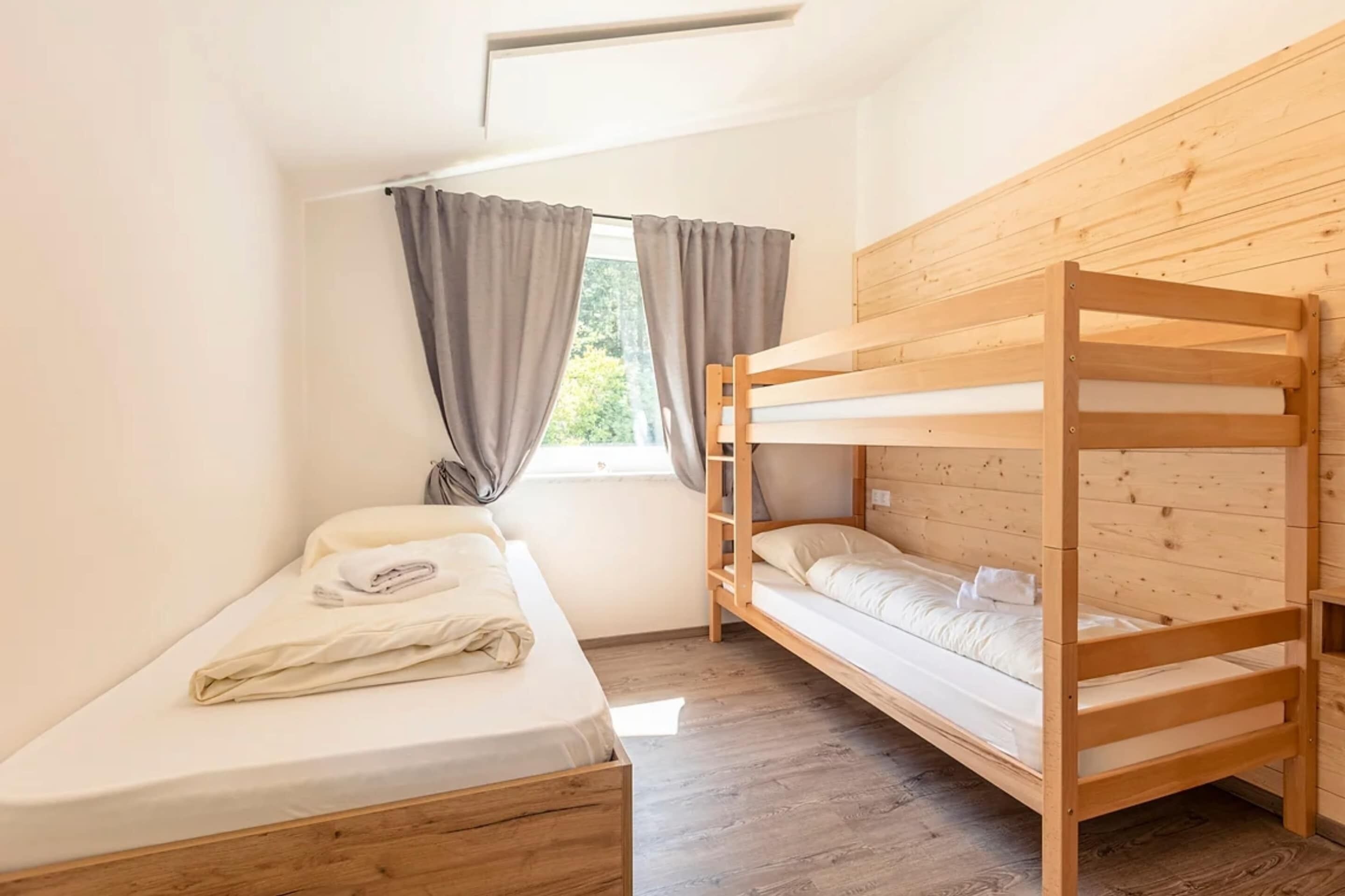 Una habitación pequeña y limpia con una cama litera, una cama individual y un ventanal con cortinas.