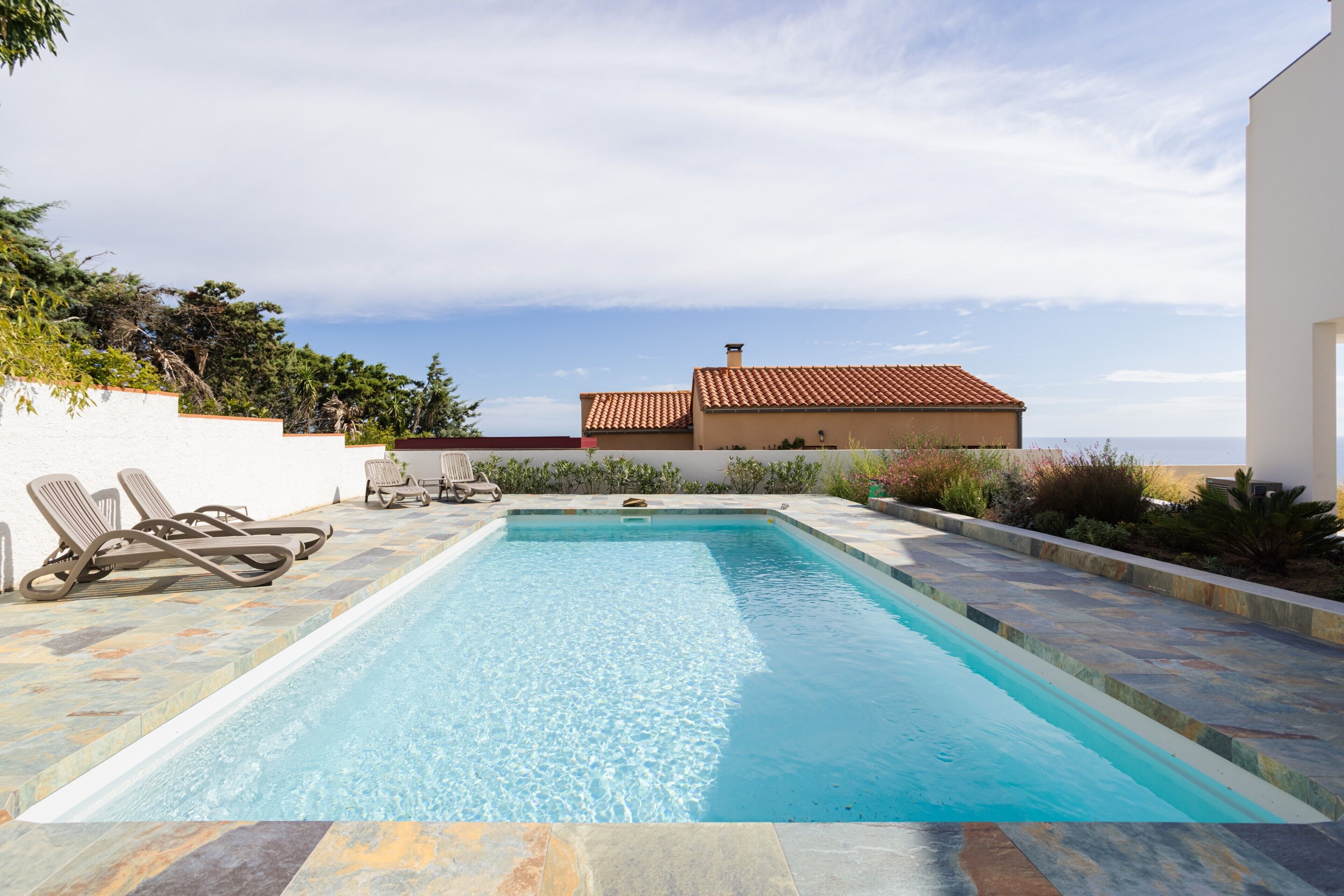 A rectangular swimming pool with a tiled deck, surrounded by a white wall and a building with a red-tiled roof.