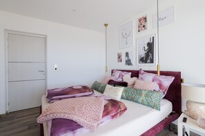 A bed with multiple pillows and a blanket, a bedside table with a lamp, and a door to the left.