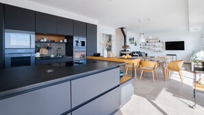 A modern kitchen with dark cabinetry, a central island, and a dining area with wooden chairs and a table. - Dream Vacation in a Beautiful Villa with Exceptional Sea View (Cerbère)