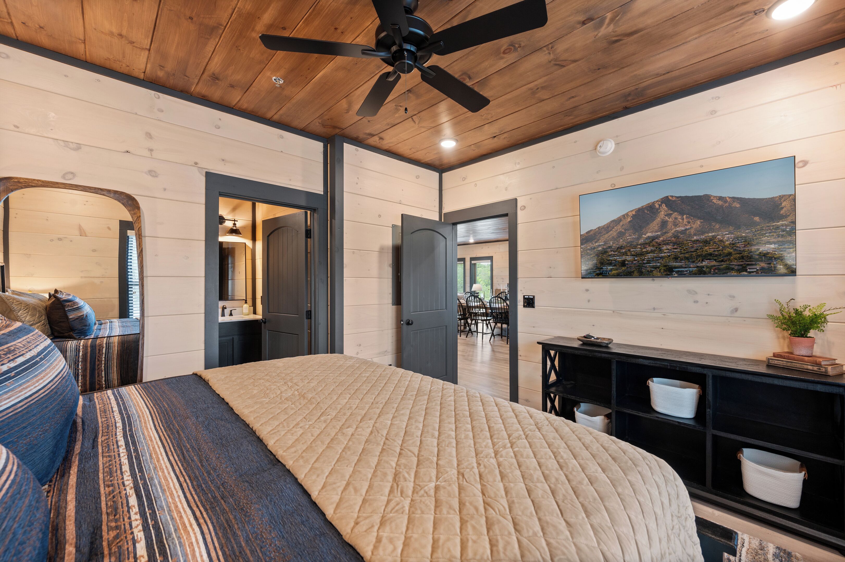 A bedroom with a large bed, a ceiling fan, a painting of a mountain landscape, and a wooden ceiling.