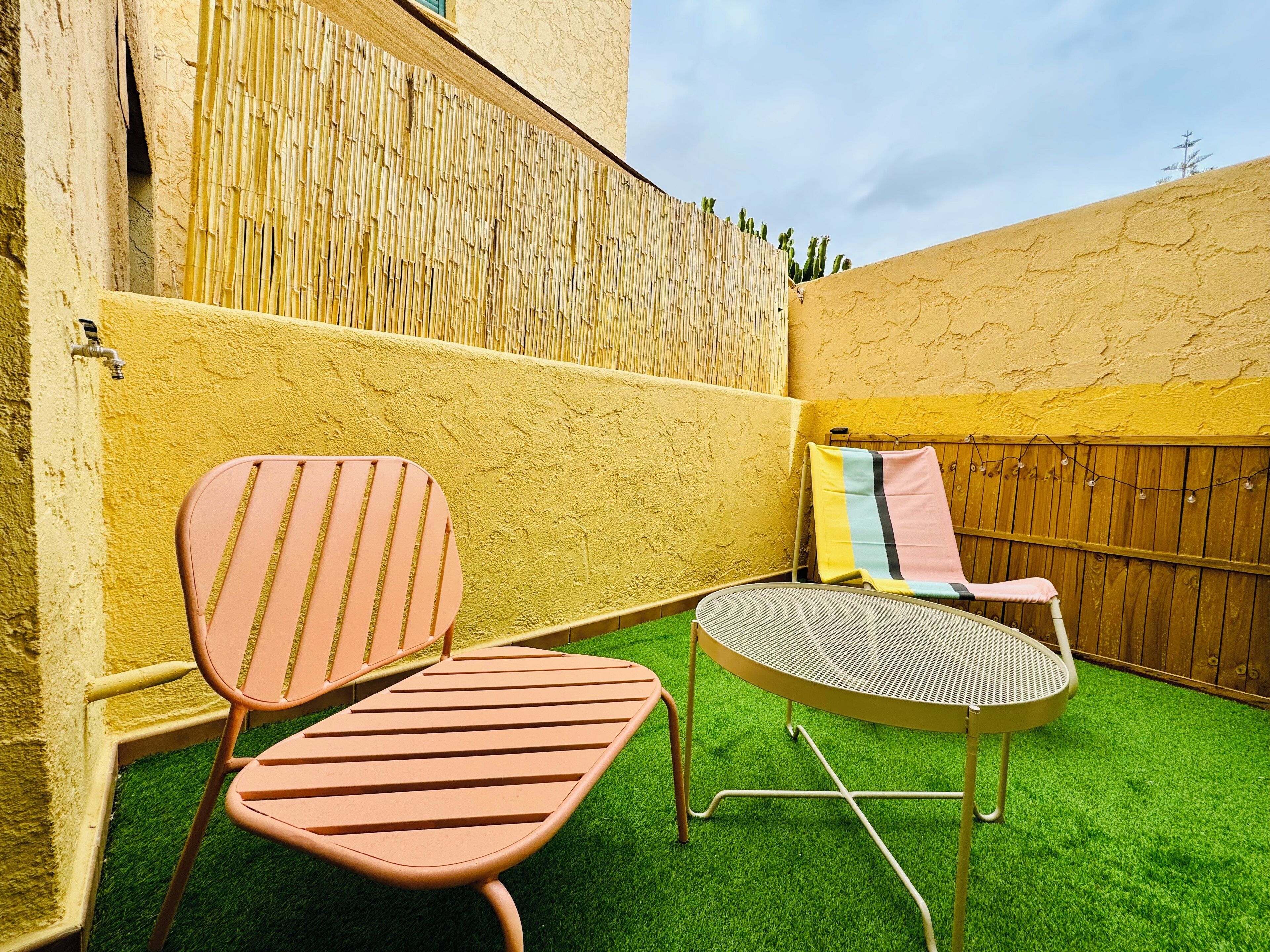 Pequeña zona ajardinada con dos sillas y una mesa redonda, rodeada por un muro amarillo y una valla de bambú.