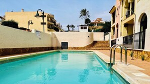 A clear swimming pool with a stone border, surrounded by buildings with balconies and a palm tree in the background. - AQUAHOME- Beautiful apartment with pool at 3 minutes walking distance from Vera Playa (Palomares)