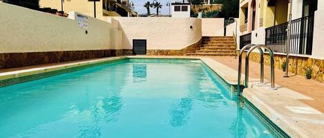 A clear swimming pool with a stone border, surrounded by buildings with balconies and a palm tree in the background.