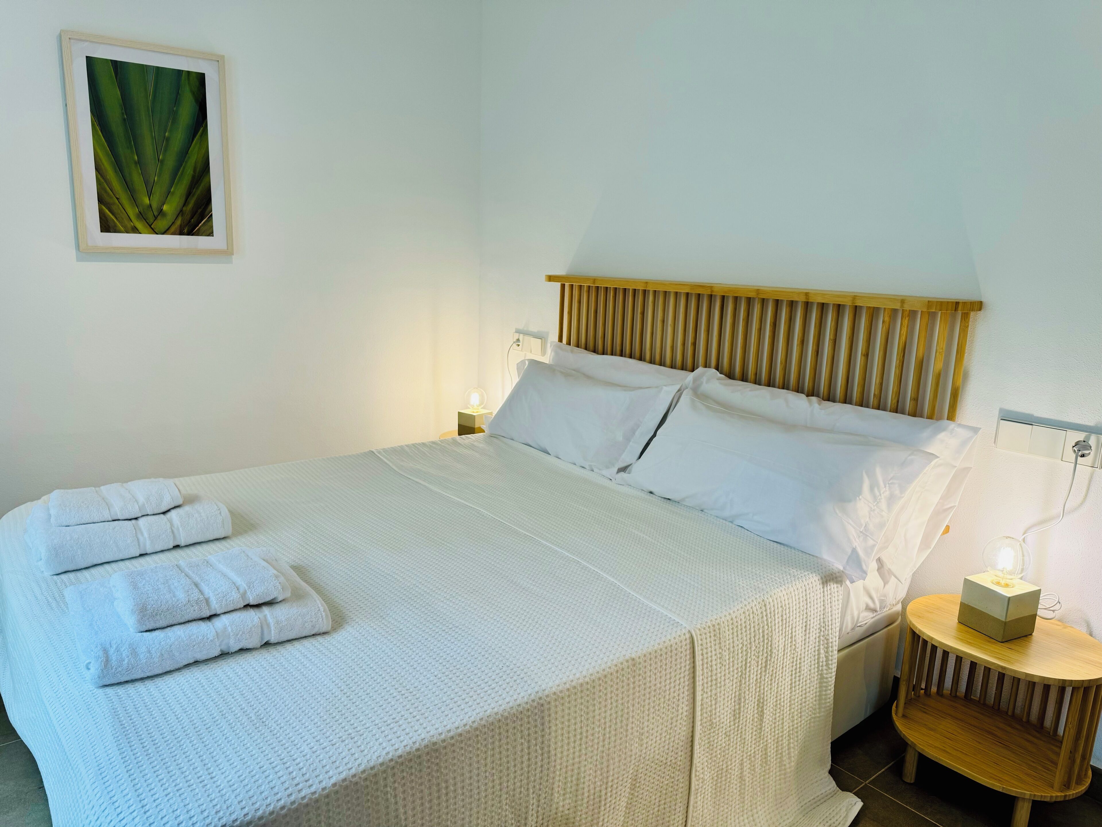 A neatly made bed with white linens and folded towels, a bedside lamp, and a small wooden nightstand with a box on it.