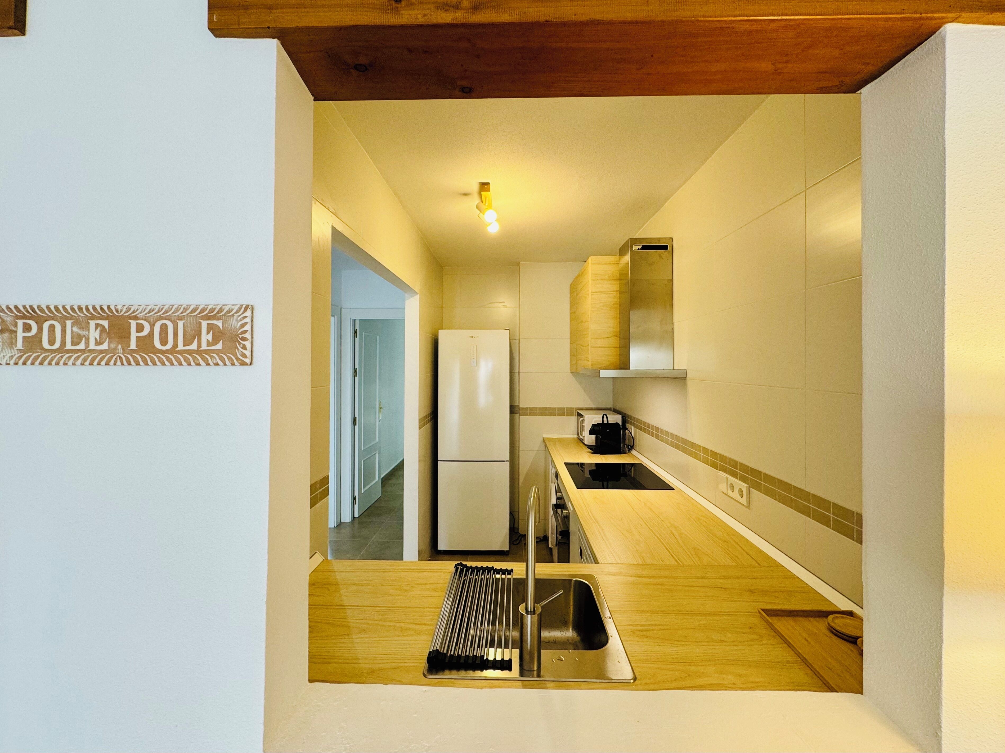 Cocina compacta con refrigerador, microondas y encimera de madera. Se ve un fregadero con un escurreplatos y un letrero que dice 