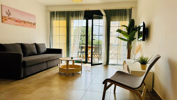 A living room with a sofa, a round table, a chair, and a large window with curtains. - AQUAHOME- Beautiful apartment with pool at 3 minutes walking distance from Vera Playa (Palomares)