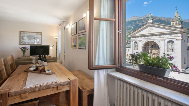 Un comedor con una mesa de madera, sillas, un televisor y una ventana que ofrece vistas a un edificio y montañas.