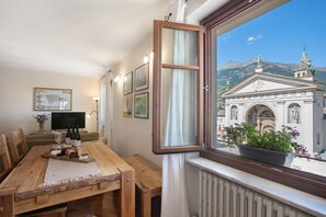 Un coin repas avec une table en bois, des chaises, un téléviseur et une fenêtre donnant sur un immeuble et des montagnes.
