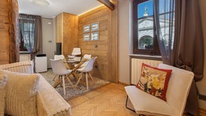 Een knus ingerichte kamer met houten details, een eethoek met een tafel en stoelen, een bank en een raam met uitzicht op een gebouw.
