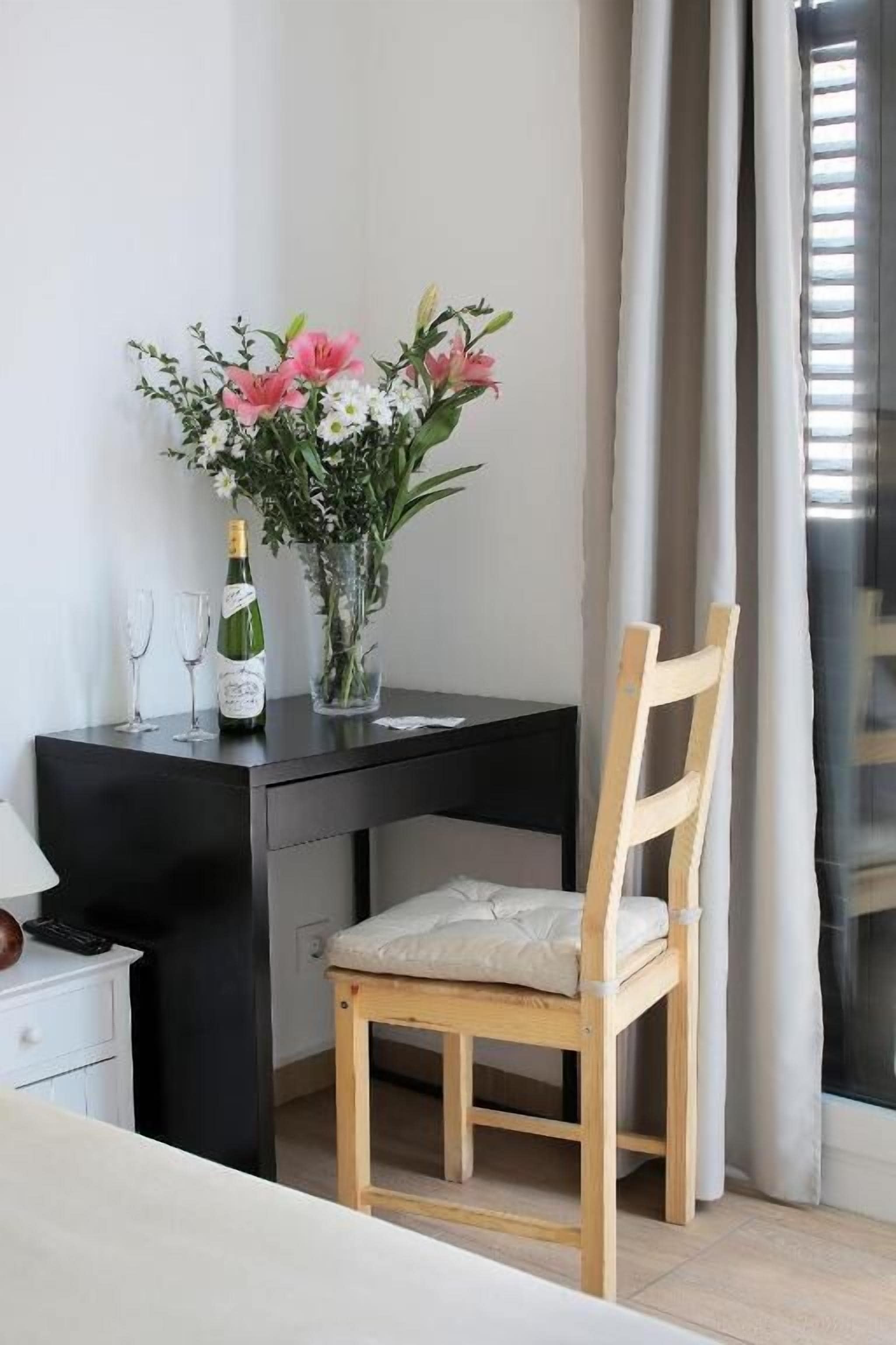 A small, modern room with a wooden chair, a desk with a vase of flowers, and a bottle of champagne.