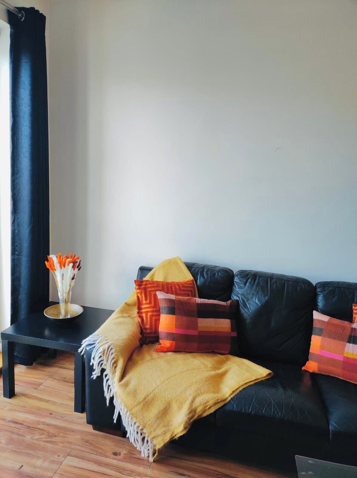 Ein schwarzes Ledersofa mit gelber Decke und bunten Kissen, ein kleiner schwarzer Nachttisch mit einer Glasvase und Holzboden.