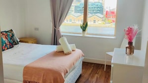 Una camera da letto con un letto, una finestra vista edifici, una pianta sul davanzale e un piccolo tavolo bianco.