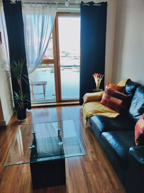 A modern living room with a glass coffee table, a leather sofa, a potted plant, and a view of a body of water through a large window. - Waterfront Views 2 Beds / 2 Baths (Dublin)