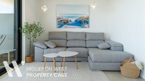 A modern living room with a grey sofa, two small white tables, a large painting of a coastal scene, a potted plant, and a woven basket.