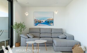 Un salon moderne avec un canapé gris, deux petites tables blanches, un grand tableau représentant un paysage côtier, une plante en pot et un panier en osier.