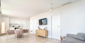 Una sala de estar moderna con un sofá, un comedor con mesa y sillas, un televisor montado en la pared y una cocina con armarios y encimera.