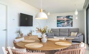 Ein modernes Wohnzimmer mit einem runden Holzes Esstisch, einem grauen Sofa und einem großen Gemälde mit Meereslandschaft.
