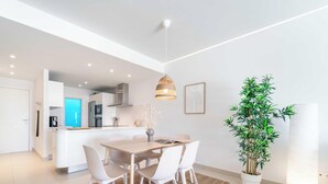 A modern kitchen with white cabinets, a dining area with a wooden table and white chairs, and a potted plant. - The Sunset Vista – West-Facing Mountain & Valley Views with Pool | Carvajal, Fuengirola (Benalmádena)