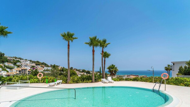 A swimming pool with lounge chairs and palm trees, surrounded by a white tiled area and a view of a coastal town. - The Sunset Vista – West-Facing Mountain & Valley Views with Pool | Carvajal, Fuengirola (Benalmádena)