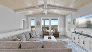A spacious living room with a high ceiling, a large sofa, two armchairs, a coffee table, and a TV. - 5513 E Pelican Drive 5 bedrooms, private pool, and easy beach access. (Oak Island)