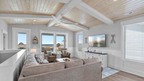 A modern living room with a large sectional sofa, a dining area with a white console table, and a view of the ocean through large windows. - 5513 E Pelican Drive 5 bedrooms, private pool, and easy beach access. (Oak Island)