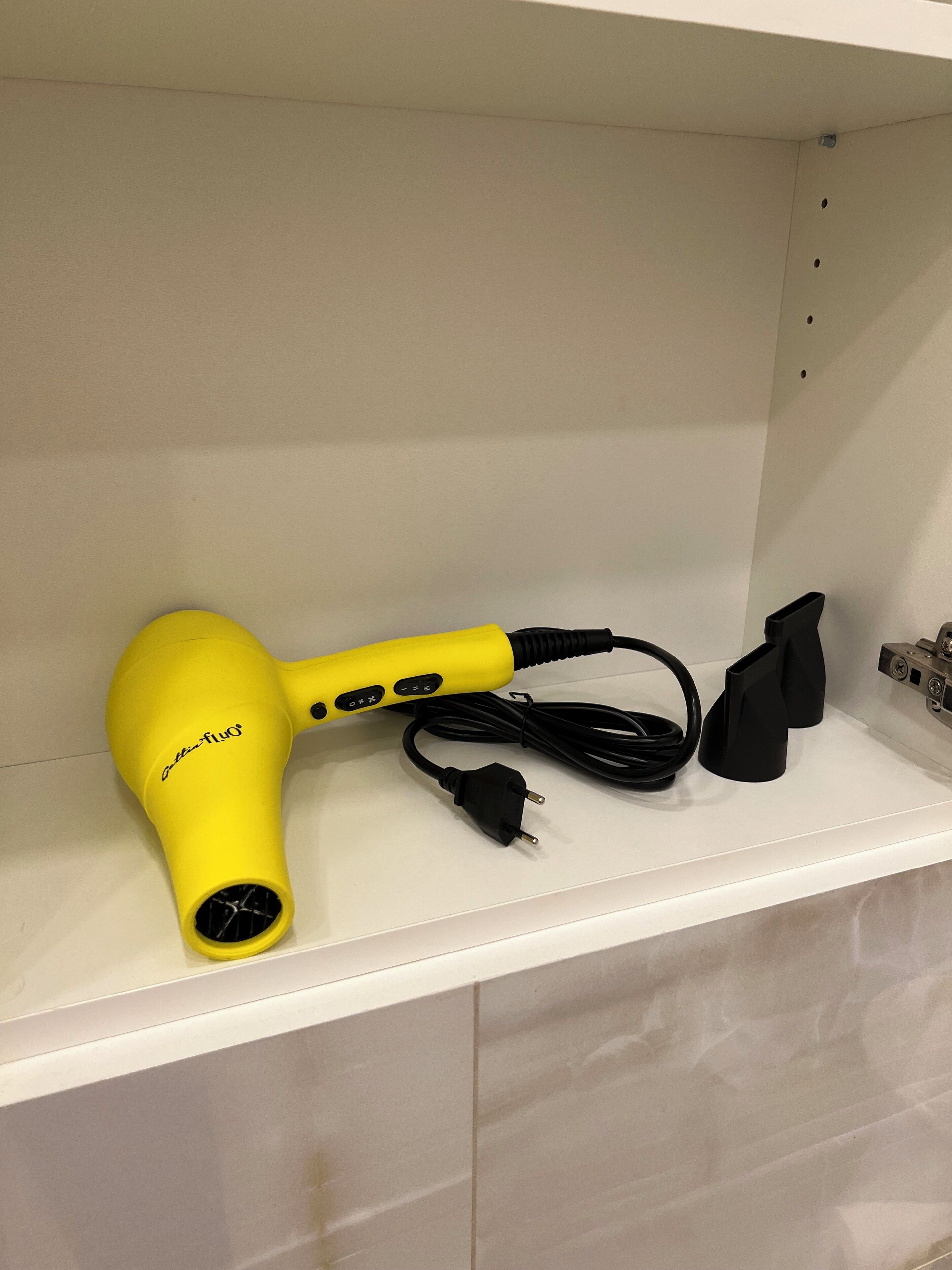 Ein gelber Fön mit Kabel und schwarzem Stecker auf einem weißen Regal.