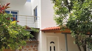 A white building with a stone wall and a door. There is a balcony with a railing and a tree in the foreground. - Olympia’s Apartment – Spacious Retreat for Up to 6 Guests (Fishta)