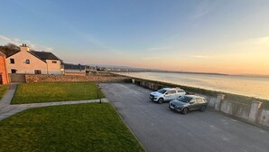 A parking area with two cars, a grassy lawn, and a building with a sloped roof. - Dolphin Bay - Beautiful 1 Bedroom Apartment (Ardersier)