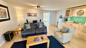 A compact living space with a sofa, a chair, a coffee table, a dining table, and a kitchenette. - Dolphin Bay - Beautiful 1 Bedroom Apartment (Ardersier)