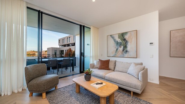 A modern living room with a sofa, armchair, and coffee table, a large abstract painting, and a view of a building through glass doors. - Signature 81 in Brighton (Brighton)
