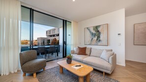 A modern living room with a sofa, armchair, and coffee table, a large abstract painting, and a view of a building through glass doors. - Signature 81 in Brighton (Brighton)