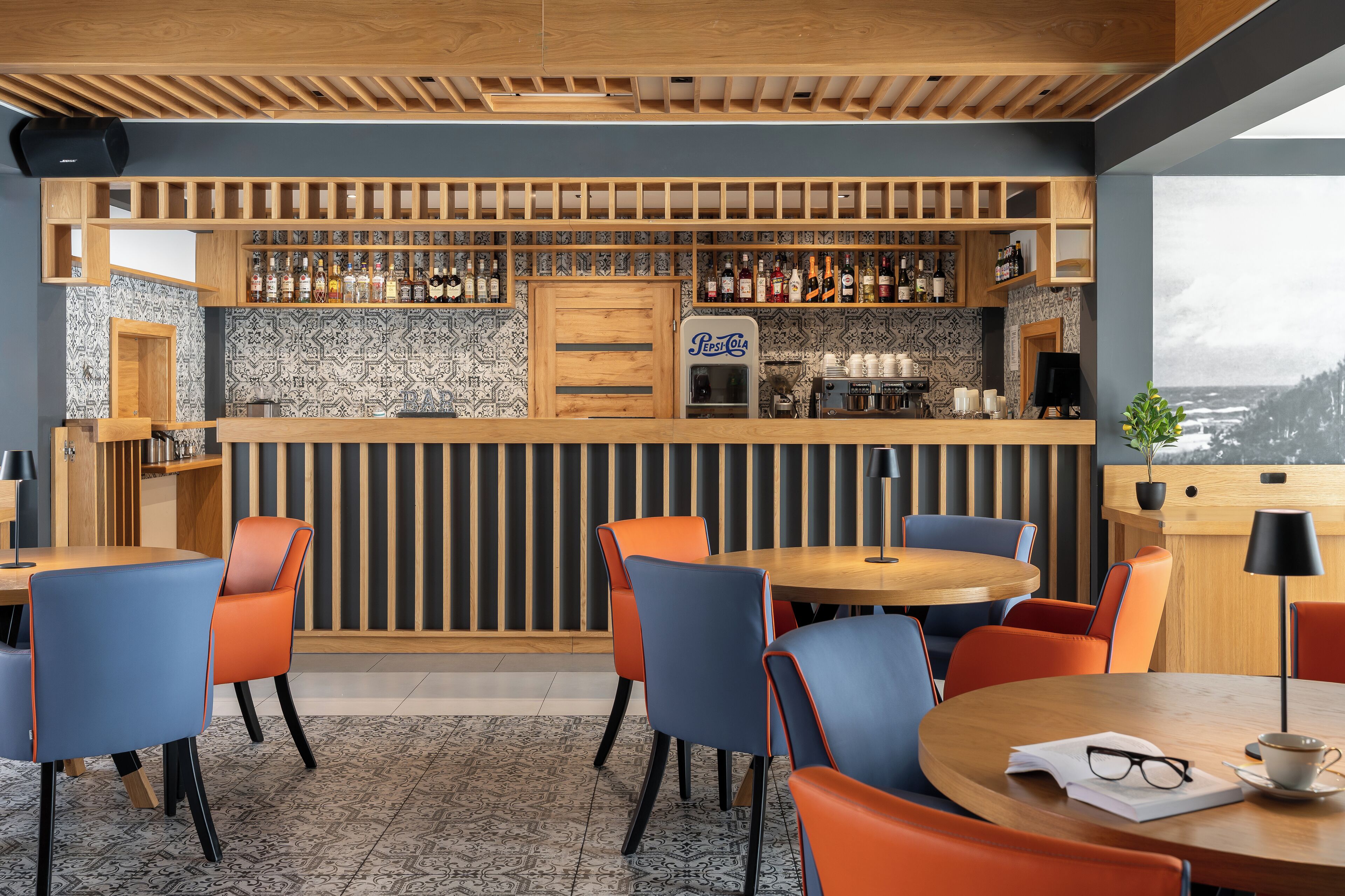A bar area with a wooden ceiling, patterned wall tiles, and a variety of bottles. There are multiple seating areas with chairs and tables.