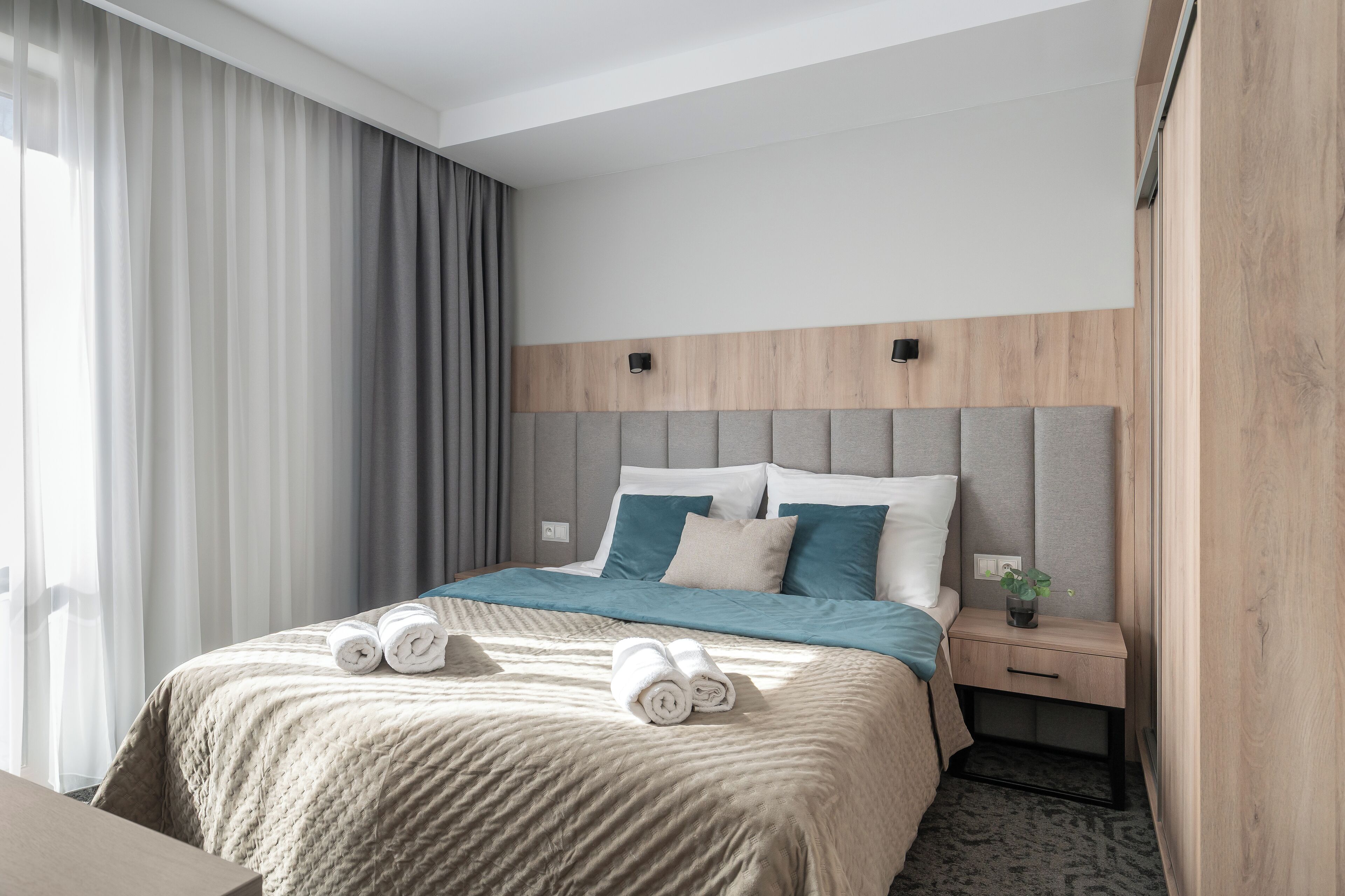 A modern hotel room with a large bed, grey curtains, a wooden headboard, and a bedside table with a plant.