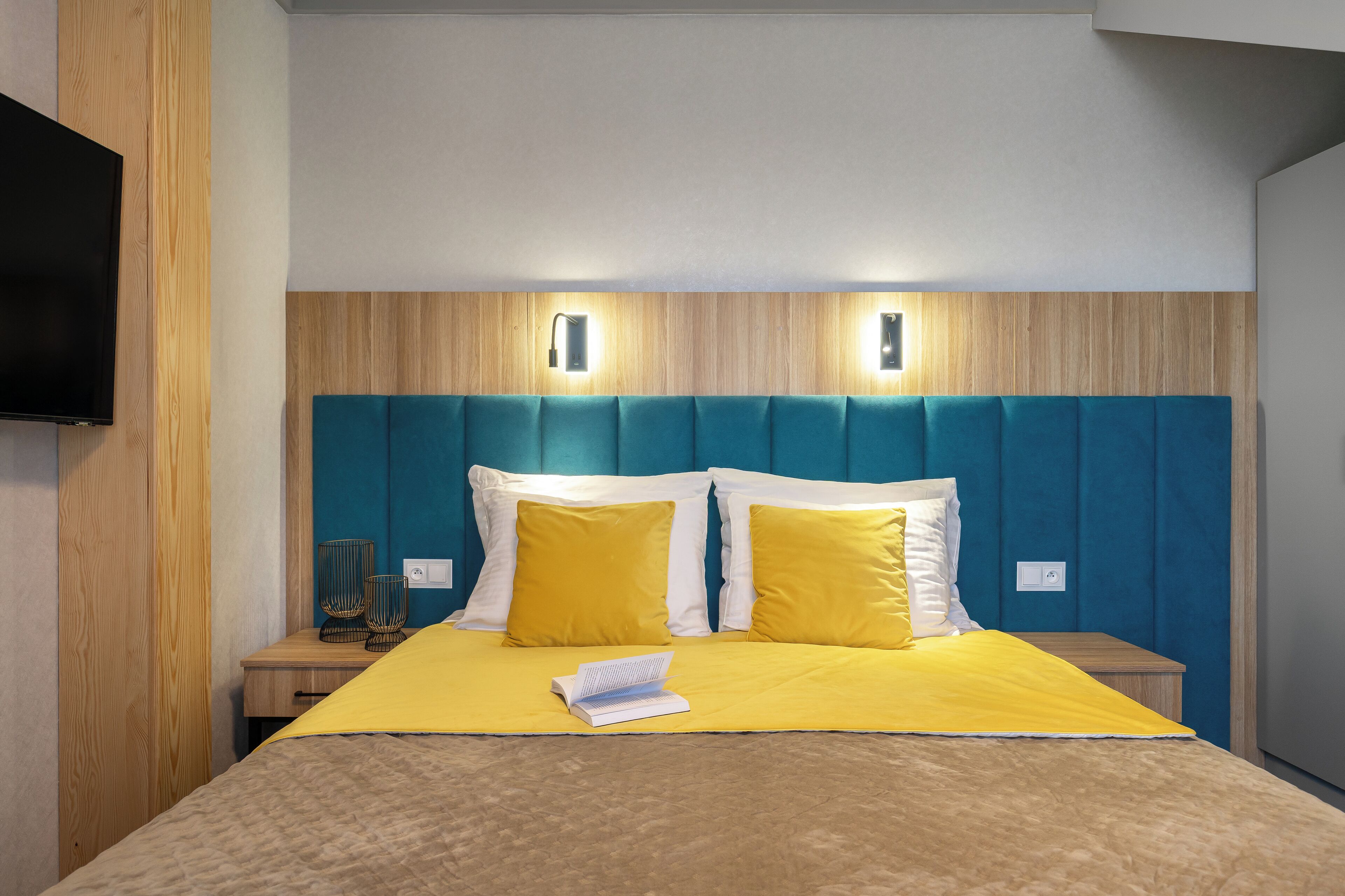 Una cama bien hecha con una colcha amarilla, dos almohadas y un libro encima. Se observan dos mesitas de noche con lámparas, un cabecero de madera y un televisor montado en la pared.