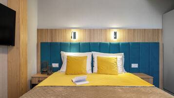 Una cama bien hecha con una colcha amarilla, dos almohadas y un libro encima. Se observan dos mesitas de noche con lámparas, un cabecero de madera y un televisor montado en la pared.